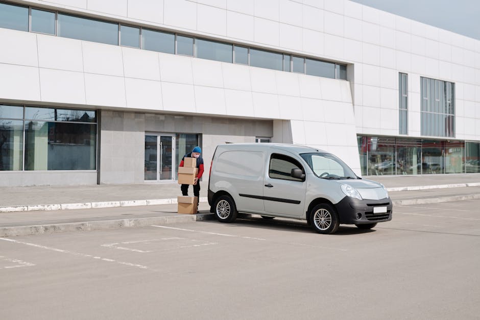 A person wearing a blue hat and red sleeves is seen standing next to a silver small cargo van parked on an empty street outside a modern white building with large glass windows. The individual is holding three cardboard boxes, one in their hands and two stacked on the ground, which appear to be part of a home relocation or furniture transport process. The van, positioned near the building's entrance, is ready for loading or unloading. The scene captures a typical outdoor setting for house removals, with the person preparing boxes for loading into the van using a trolley or directly. The environment includes an orderly pavement and parking area, consistent with relocation services operating within urban or suburban residential zones. Man with Van Holland Park uses these logistics for efficient packing, loading, and moving within the local area, adhering to parking and loading rules for removals vans close to the property.