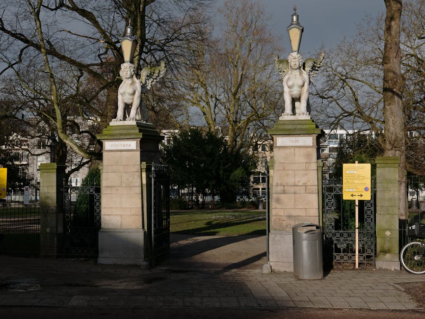 The image shows the entrance gate to a park with two stone pillars, each topped with a decorative sculpture of a mythical creature with wings, sitting beneath street lamps. The sculptures are positioned on square pedestals inscribed with the words 'WETHEIM' and 'PARK'. The gate is partially open, revealing a pathway leading into a green park area with large, leafless trees and a backdrop of residential buildings. To the right of the gate, there is a yellow information sign and a grey trash bin placed on the paved sidewalk, with a bicycle wheel visible partially on the far right. The scene is illuminated by daylight, and the surrounding environment suggests an urban setting suitable for home relocation or furniture transport services, as offered by Man with Van Holland Park.