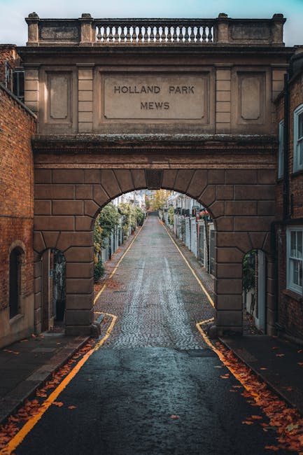 A view of an open wrought iron gate leading into a park area with tall, leafy green trees and a pathway beyond. The ground in the foreground is wet, creating a large puddle that reflects the trees, gate, and parts of the surrounding environment, including nearby buildings in the distance. The scene is well-lit with natural daylight, and there are benches and lampposts visible along the pathway. This setting illustrates the outdoor environment for a house relocation process, where furniture and boxes from Man with Van Holland Park might be moved through the gate into or out of the park area for home relocation or furniture transport purposes.