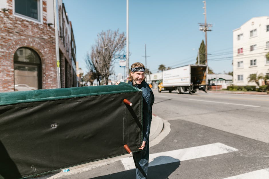 A man with long hair, wearing a headband and casual clothing, is seen outdoors on a street corner during daylight hours. He is smiling and carrying a large, rectangular piece of furniture or equipment covered in black fabric with brown handles, possibly part of home relocation or furniture transport. In the background, there is a white truck with an open cargo area on the road, and the scene includes a brick building with arched windows on the left, a modern white multi-story building on the right, and trees without leaves. The street features a crosswalk, power lines, and a traffic sign, indicating an urban environment suitable for moving and delivery services. The scene is well-lit, with shadows cast on the pavement, and appears to be part of a loading process or park-and-load scenario connected to professional removals by Man with Van Holland Park as detailed on the provided guide page.
