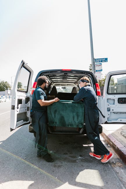 Two movers from Man with Van Holland Park are engaged in a home relocation task, lifting and carefully handling a large, dark green upholstered piece of furniture wrapped in protective padding to prevent damage during transportation. They are positioned at the back of a white van with open rear doors, inside which various packed items are visible, indicating an active packing and loading process. One worker, wearing a navy blue uniform and glasses, is on the right, pulling the furniture towards the van, while the other, in a dark shirt and trousers, supports from the left. The loading area is on a paved street next to a sidewalk with a street sign visible overhead, under bright daylight with clear skies. The scene highlights the careful logistics involved in furniture transport and moving services, emphasizing the professional approach of [COMPANY_NAME] in managing house removals with proper equipment like blankets and straps for secure transit during shifting of household belongings in the context of detailed packing and moving operations referenced in the Holland Walk logistics guide.