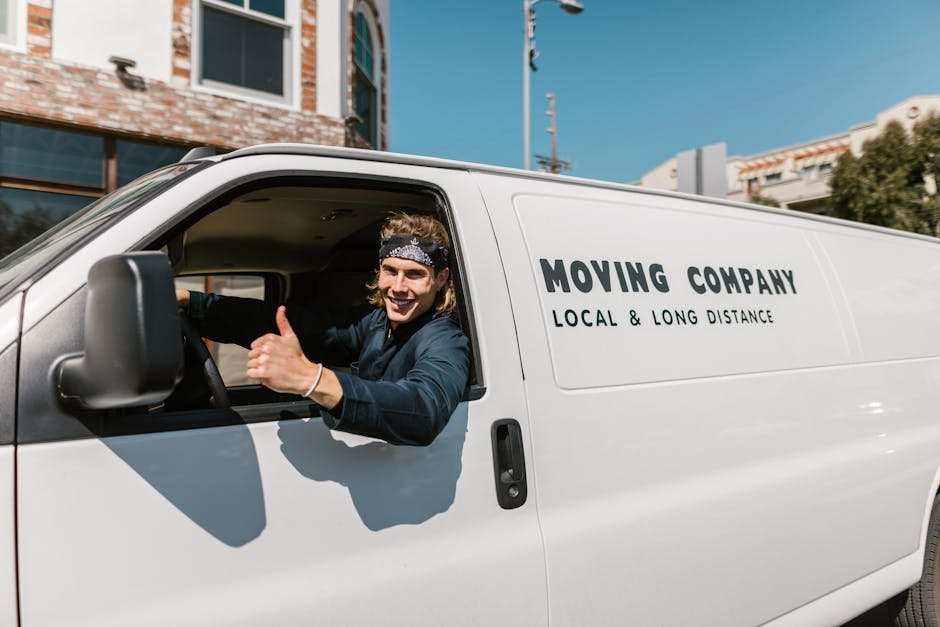 A young man sitting in the driver's seat of a white moving van branded with the words 'MOVING COMPANY' and 'LOCAL & LONG DISTANCE', inside an urban residential area with clear blue skies. The man is smiling, wearing a denim shirt and a patterned headband, and is giving a thumbs-up gesture towards the camera. The open window reveals the vehicle's interior, and the van is parked on a street with modern buildings and greenery in the background. Visible inside the van are cardboard boxes and packing materials, indicating a home relocation or furniture transport process. Occasionally, Man with Van Holland Park's professional team is involved in loading or preparing household items for transfer, with equipment such as trolleys or blankets supporting the moving operations.