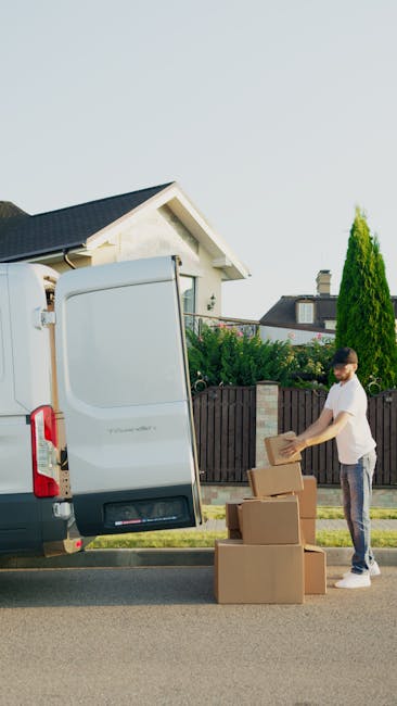 A man wearing a white t-shirt and jeans is standing on the pavement next to an open white cargo van, part of a home relocation process managed by Man with Van Holland Park. He is handling a cardboard box, placing or removing it from a stack of several boxes of various sizes, which are packed with packing materials such as brown paper. The van is parked close to a residential property with a dark tiled roof, white walls, and a small front yard separated by a wooden fence. In the background, there are greenery including a tall coniferous tree and other bushes, with neighboring houses visible above the fence and a clear, bright sky overhead. The loading area is outside, with the open vehicle door revealing the rear compartment used for furniture transport and packaging during the packing and moving process, illustrating the typical steps involved in house removals and furniture transport services provided by Man with Van Holland Park.