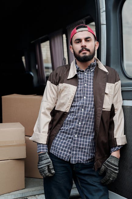 A young man sitting in the driver's seat of a white moving van branded with the words 'MOVING COMPANY' and 'LOCAL & LONG DISTANCE', inside an urban residential area with clear blue skies. The man is smiling, wearing a denim shirt and a patterned headband, and is giving a thumbs-up gesture towards the camera. The open window reveals the vehicle's interior, and the van is parked on a street with modern buildings and greenery in the background. Visible inside the van are cardboard boxes and packing materials, indicating a home relocation or furniture transport process. Occasionally, Man with Van Holland Park's professional team is involved in loading or preparing household items for transfer, with equipment such as trolleys or blankets supporting the moving operations.