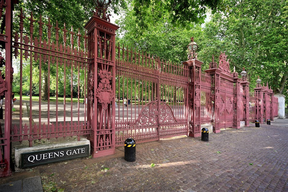 A large, ornate, pink-painted wrought iron gate with decorative finials and detailed panels, situated at Queens Gate entrance. The gate is supported by tall, matching pillars topped with classic-style lanterns. The gate is partly open, revealing a pathway and a park area beyond with green trees and a paved walkway. The ground in front of the gate is cobblestone, with a black and yellow bollard in place. To the left of the gate, a black sign with white letters reads 'QUEENS GATE.' The background includes lush foliage from mature trees with dense green leaves, providing a shaded environment indicative of an urban park setting, suitable for house removal access or furniture transport as part of a home relocation service by Man with Van Holland Park.