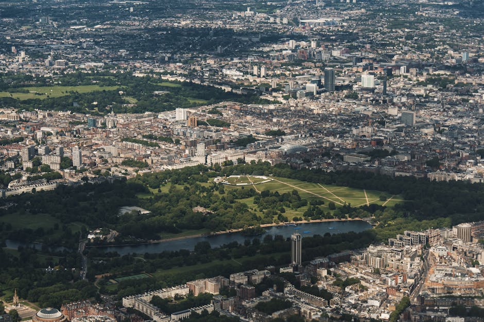 An aerial view of Kensington Gardens in the Royal Borough of Kensington and Chelsea, showcasing a large rectangular green park with pathways crossing through it, surrounded by dense urban buildings and streets. In the foreground, a river runs along the edge of the park, with trees lining its banks. The scene is captured during daylight hours with clear visibility of the cityscape extending into the distance. This image highlights the urban environment that house removal services by Man with Van Holland Park may operate within, emphasizing the need for careful packing and transportation of furniture and belongings during home relocation or furniture transport in this area.